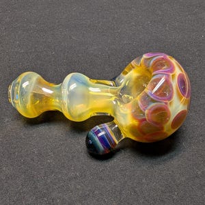 May include: A hand-blown glass pipe with a yellow, orange, and purple color scheme. The pipe features a rounded bowl with purple polka dots, a cylindrical stem, and a small, rounded mouthpiece. The glass has a glossy finish.