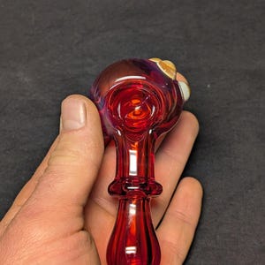 May include: A hand-blown glass pipe with a vibrant red hue, transitioning to a deep purple and gold at the bowl. The pipe features a bulbous bowl and a flared, rounded base. The glass is smooth and reflective.
