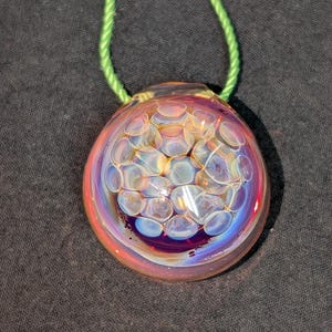 May include: A round, iridescent glass pendant with a bubble-like design, featuring shades of pink, purple, and blue. The pendant is suspended from a green cord, creating a unique and eye-catching accessory.