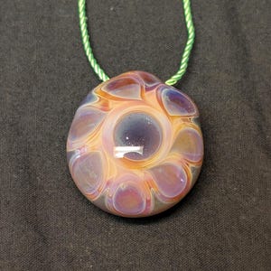 May include: A glass pendant necklace with a distinctive, swirling pattern. The pendant has a dark blue centre, encircled by lighter orange and purple shades. It is suspended from a green cord, providing a vivid contrast.
