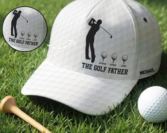 May include: A white golf hat with a golf ball pattern, the text "THE GOLF FATHER", and a silhouette of a golfer. A golf ball and a wooden tee are on the green grass. The hat has the name "MICHAEL" on the side.