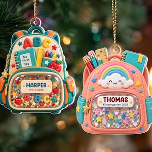 May include: Two backpack-shaped ornaments hanging by gold cords. The left one is teal and cream with 'HARPER Pre-K 2026' and 'ABC' text. The right one is peach and teal with a rainbow, 'THOMAS Kindergarten 2026' text, and shaker-style glitter.