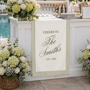 May include: A white bar runner featuring a sage green striped border and the text Cheers to The Smiths EST. 2026 in a dark green script and serif font. The runner is displayed on a white bar counter decorated with large white and green floral arrangements.