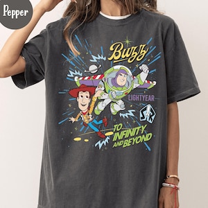 May include: Dark gray t-shirt featuring a graphic of Buzz Lightyear and Woody from Toy Story. The design includes the text "Buzz Lightyear" and "To Infinity and Beyond" in a colorful, cartoon style. The shirt has a relaxed fit.