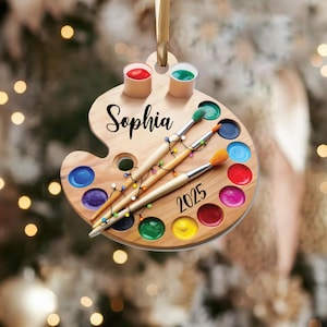 May include: A wooden artist's palette Christmas ornament with the name "Sophia" and the year "2025" in black script. The palette features paint colors, paintbrushes, and small paint cups. A string of colorful lights is wrapped around the paintbrushes.