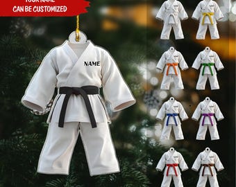 May include: White karate uniform ornament with a black belt and the word "NAME" printed on the chest. The ornament hangs from a gold string. Additional ornaments with colored belts are also shown.