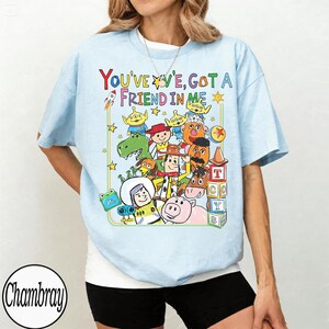 May include: Pale blue t-shirt with a vibrant cartoon graphic and the words "YOU'VE GOT A FRIEND IN ME". The design includes a rocket, stars, and various animated figures. The shirt has short sleeves.
