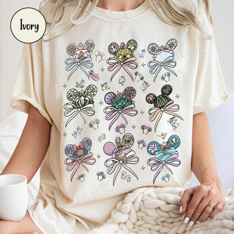 May include: Ivory-colored t-shirt featuring colorful illustrations of Mickey Mouse ears with bows and character-themed designs. The shirt has a relaxed fit and short sleeves. The word "Ivory" is in a circle in the upper left corner.