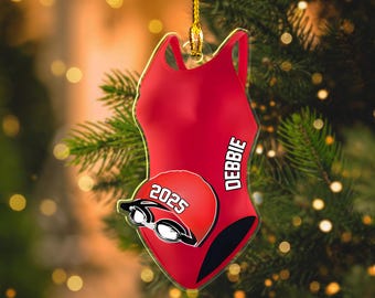 May include: A red swimsuit-shaped ornament with the name "DEBBIE" printed vertically. A red swim cap with the year "2025" and swim goggles is on the front. The ornament hangs from a gold string against a blurred Christmas tree background.