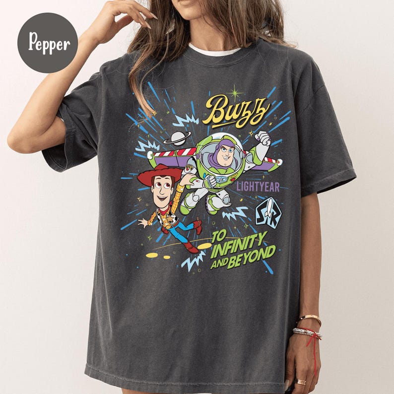 May include: Dark gray t-shirt featuring a graphic of Buzz Lightyear and Woody from Toy Story. The design includes the text "Buzz Lightyear" and "To Infinity and Beyond" in a colorful, cartoon style. The shirt has a relaxed fit.