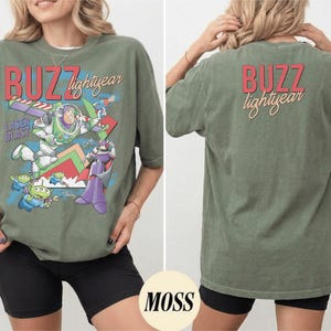 May include: Olive green t-shirt featuring a graphic of Buzz Lightyear and other characters from the movie. The front of the shirt has the words "BUZZ lightyear" in red and yellow. The back of the shirt also has the words "BUZZ lightyear".