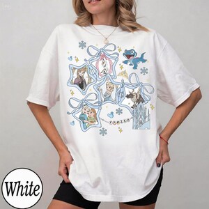 Może przedstawiać: Biała koszulka z grafiką przedstawiającą postacie z filmu "Kraina Lodu". Projekt zawiera obrazy Elsy, Anny, Olafa, Kristoffa i Svena, obramowane niebieskimi wstążkami i płatkami śniegu. Wyświetlane jest r&oacute;wnież słowo "FROZEN".