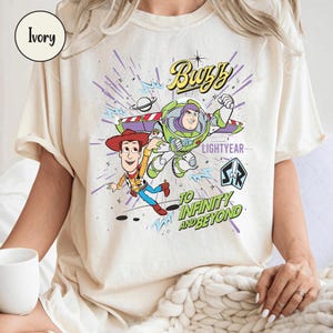 May include: Ivory-colored t-shirt featuring a graphic of Buzz Lightyear and Woody from Toy Story. The design includes the text "Buzz Lightyear" and "To Infinity and Beyond" in a playful font. The shirt has short sleeves.