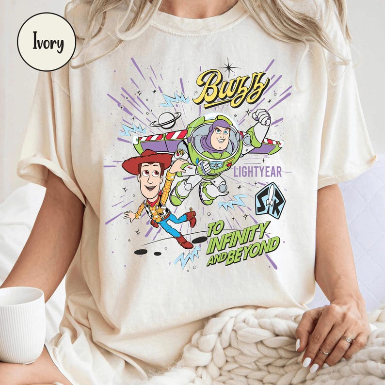 May include: Ivory-colored t-shirt featuring a graphic of Buzz Lightyear and Woody from Toy Story. The design includes the text "Buzz Lightyear" and "To Infinity and Beyond" in a playful font. The shirt has short sleeves.