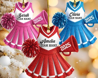 May include: Three cheerleader ornaments in pink, red, and blue, each with a matching pom-pom and megaphone. The ornaments feature names and "TEAM NAME" text, with the year "2025" on the megaphones. The ornaments are hanging from gold strings.