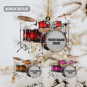 May include: Acrylic 2D flat drum set ornament with the text "YOUR NAME 2025" on the bass drum. The ornament is available in red, orange, and pink. The ornament is hanging from a gold string.