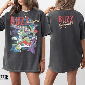 May include: Dark gray t-shirt featuring a graphic of Buzz Lightyear with the text "BUZZ lightyear" in red and yellow. The front of the shirt has a colorful design with the words "LASER BLAST". The back of the shirt also has the text "BUZZ lightyear".