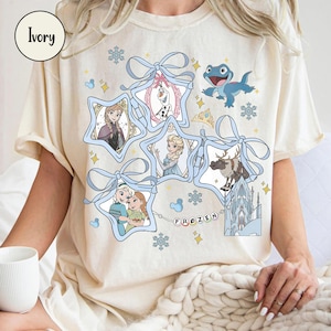 Może przedstawiać: Kremowa koszulka z grafiką z filmu "Kraina Lodu". Projekt przedstawia postacie takie jak Anna, Elsa, Olaf, Kristoff i Sven, w ramkach w kształcie gwiazd, połączonych niebieskimi wstążkami i płatkami śniegu. Słowo "FROZEN" jest napisane kolorowymi literami.