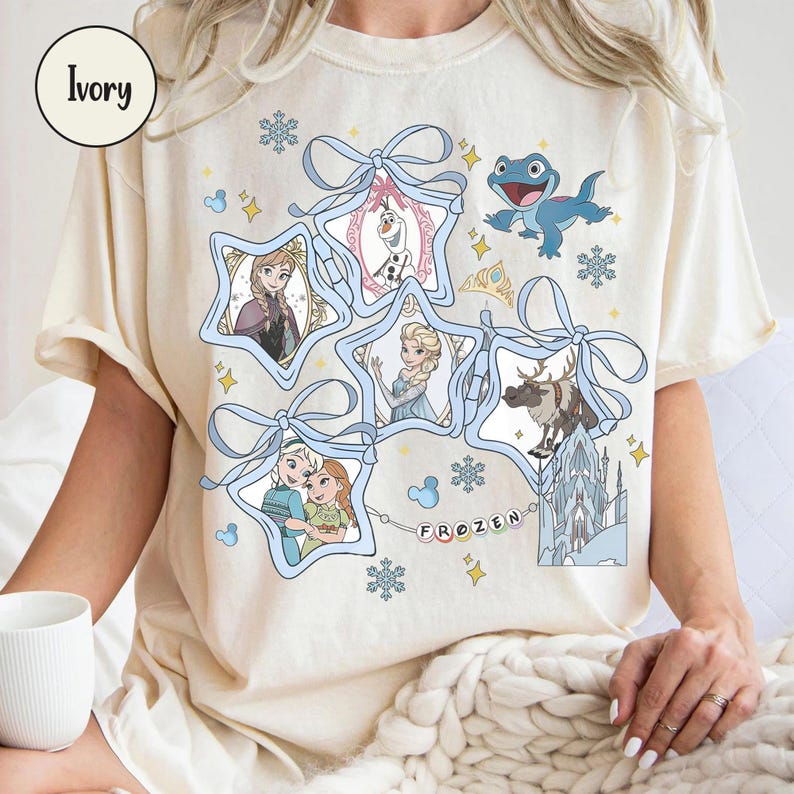 Może przedstawiać: Kremowa koszulka z grafiką z filmu "Kraina Lodu". Projekt przedstawia postacie takie jak Anna, Elsa, Olaf, Kristoff i Sven, w ramkach w kształcie gwiazd, połączonych niebieskimi wstążkami i płatkami śniegu. Słowo "FROZEN" jest napisane kolorowymi literami.