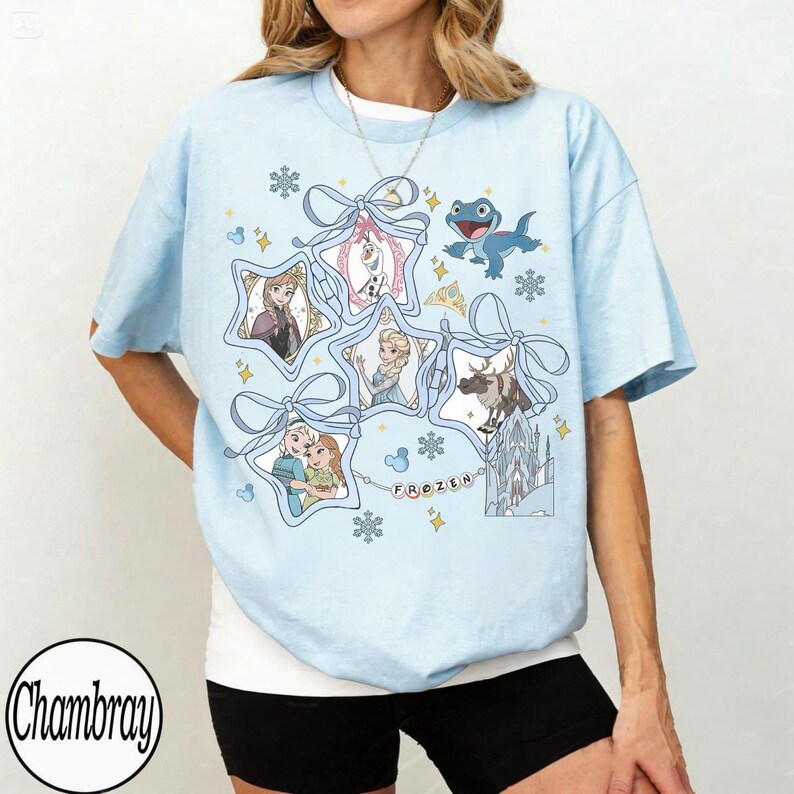 Może przedstawiać: Jasnoniebieska koszulka z motywem z filmu "Kraina Lodu". Projekt zawiera wizerunki postaci takich jak Elsa, Anna, Olaf i Sven, umieszczone w gwiazdowych ramkach związanych niebieskimi wstążkami. Widnieje r&oacute;wnież napis "FROZEN".