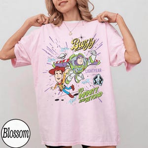 May include: Light pink t-shirt featuring Buzz Lightyear and Woody from Toy Story. The graphic includes the text "Buzz Lightyear" and "To Infinity and Beyond" in a colorful design. The shirt has short sleeves and a crew neck.