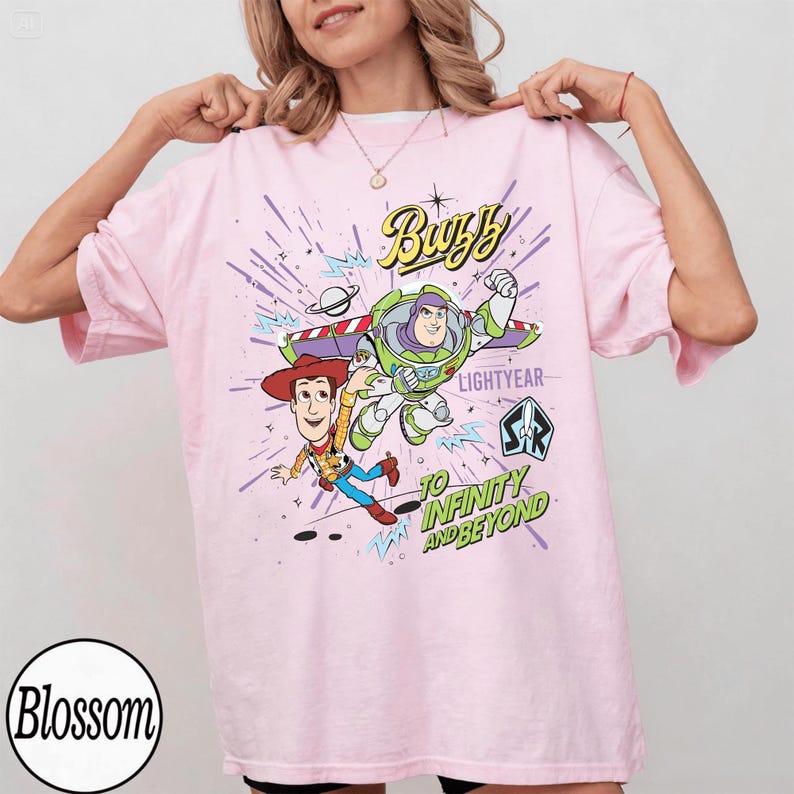 May include: Light pink t-shirt featuring Buzz Lightyear and Woody from Toy Story. The graphic includes the text "Buzz Lightyear" and "To Infinity and Beyond" in a colorful design. The shirt has short sleeves and a crew neck.