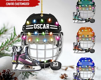 May include: A hockey helmet ornament with the name "OSCAR" and a hockey skate. The ornament has a black helmet with colorful lights and a hockey puck with the number "23". The text "NUMBER, YOUR NAME CAN BE CUSTOMIZED" is visible.