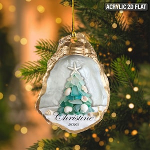 May include: Acrylic 2D flat Christmas ornament shaped like an oyster shell. The interior features a Christmas tree made of sea glass and seashells. The name "Christine" and the year "2025" are written on the bottom.