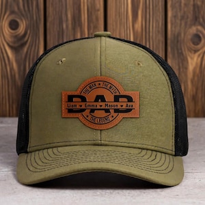 May include: Olive green and black trucker hat with a leather patch that reads "DAD" with the words "The Man, The Myth, The Legend" and the names Liam, Emma, Mason, and Ava. The hat has a curved brim.