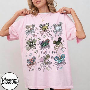 May include: A light pink t-shirt featuring a pattern of Mickey Mouse ears with decorative bows. The ears have various designs and colors, including floral, striped, and jeweled elements. The word "Blossom" is in a white circle at the bottom left.