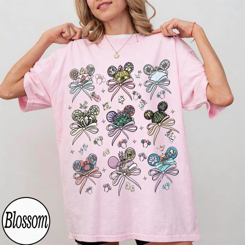 May include: A light pink t-shirt featuring a pattern of Mickey Mouse ears with decorative bows. The ears have various designs and colors, including floral, striped, and jeweled elements. The word "Blossom" is in a white circle at the bottom left.