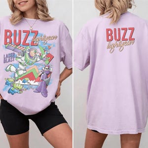 May include: A light purple t-shirt featuring a graphic of Buzz Lightyear and the text "BUZZ lightyear" in red and gold. The back of the shirt also has the text "BUZZ lightyear" in red and gold.