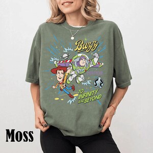 May include: Moss green t-shirt featuring a graphic of Buzz Lightyear and Woody from Toy Story. The design includes the text "Buzz Lightyear" and "To Infinity and Beyond".
