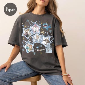 Może przedstawiać: Ciemnoszara koszulka z grafiką postaci z filmu Kraina Lodu. Projekt zawiera obrazy Elsy, Anny, Olafa, Kristoffa i zamku, ułożone w ramki w kształcie gwiazd z niebieskimi wstążkami i płatkami śniegu. Obecne jest r&oacute;wnież słowo "Frozen".