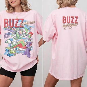 May include: A light pink t-shirt featuring a graphic of Buzz Lightyear and other characters from the movie Toy Story. The front of the shirt has the words "BUZZ lightyear" in red and yellow. The back of the shirt has the words "BUZZ lightyear" in red and yellow.