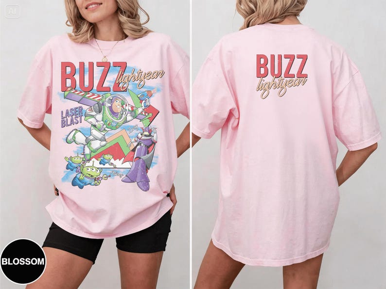 May include: A light pink t-shirt featuring a graphic of Buzz Lightyear and other characters from the movie Toy Story. The front of the shirt has the words "BUZZ lightyear" in red and yellow. The back of the shirt has the words "BUZZ lightyear" in red and yellow.