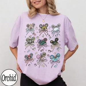 May include: A light purple t-shirt featuring a repeating pattern of Mickey Mouse ears with bows and decorative elements. The design includes various colors and styles, with the word "Orchid" in a circle at the bottom left.
