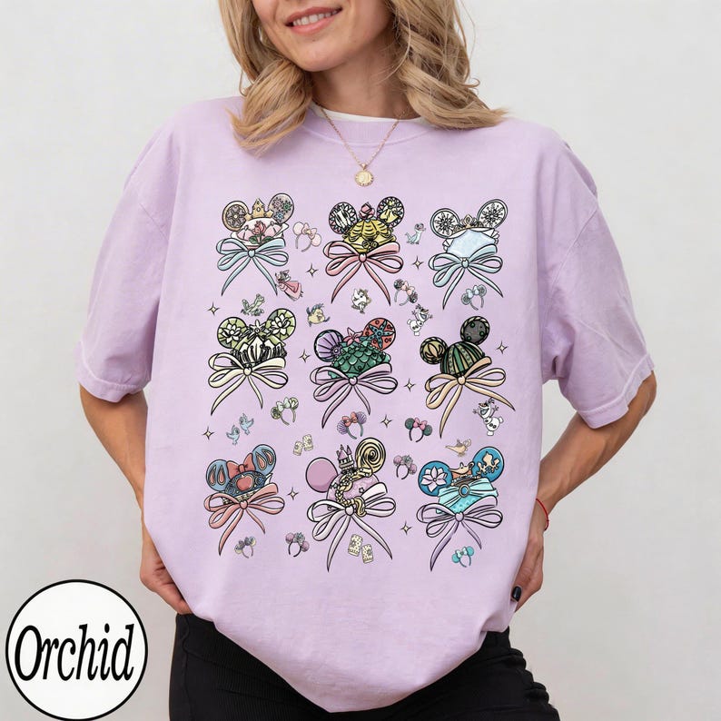 May include: A light purple t-shirt featuring a repeating pattern of Mickey Mouse ears with bows and decorative elements. The design includes various colors and styles, with the word "Orchid" in a circle at the bottom left.
