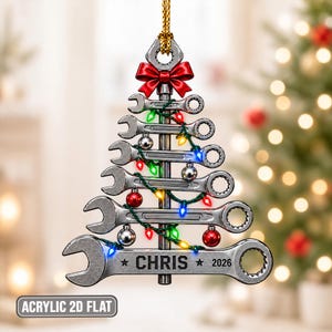 May include: A Christmas tree-shaped ornament made of stacked silver-toned wrenches, topped with a red bow and a gold hanging cord. The design features colourful string lights, small silver and red ornaments, and the text CHRIS 2026.