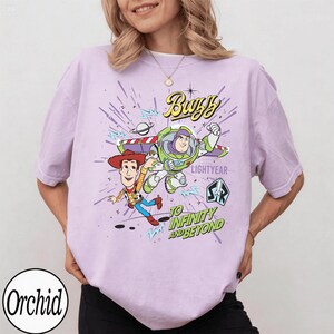 May include: A light purple t-shirt featuring a graphic of Buzz Lightyear and Woody from Toy Story. The design includes the words "Buzz Lightyear" and "To Infinity and Beyond" in a playful font. The shirt has short sleeves.