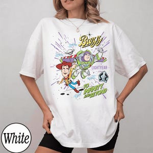 May include: White t-shirt featuring a graphic of Buzz Lightyear and Woody from Toy Story. The design includes the text "Buzz Lightyear" and "To Infinity and Beyond" in a colorful, cartoon style. The shirt has short sleeves.