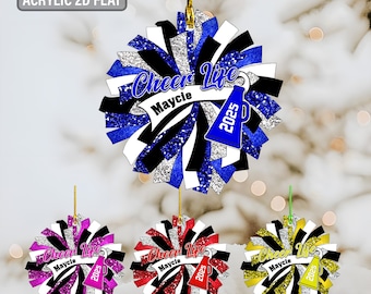 May include: Acrylic 2D flat cheerleading ornament with the text "Cheer Life Maycie 2025". The ornament is a pom-pom design with blue, black, white, and silver glitter accents. Additional ornaments in pink, red, and yellow are also shown.
