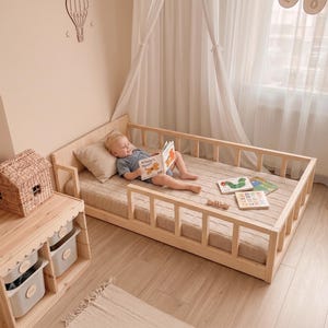 Può includere: Un letto a terra in legno a profilo ribassato con sponde di sicurezza a doghe, un materasso beige e un mobile contenitore in legno abbinato con due ripiani aperti e contenitori grigi, sormontato da un cesto intrecciato a forma di casa.