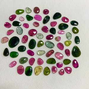 May include: A collection of 40+ green, pink, and yellow gemstones. The gemstones are various shapes and sizes, and have a natural, rough finish.