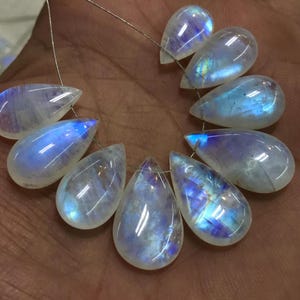 May include: A selection of teardrop-shaped moonstone beads strung on wire. The translucent stones exhibit a shimmering play of colour, with flashes of blue and iridescent hues. The beads are smooth and polished, reflecting light.