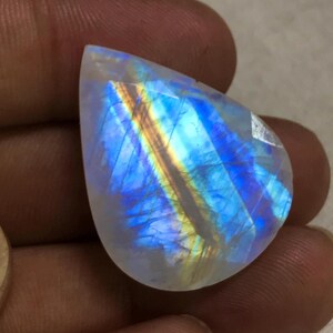 Puede incluir: Una piedra lunar facetada en forma de pera. La piedra muestra un juego de colores, con destellos de azul, amarillo y naranja dentro del material blanco lechoso translúcido. La gema se sostiene entre dos dedos.