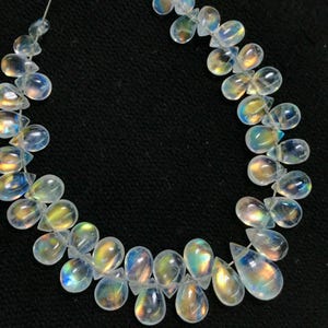 May include: A necklace of iridescent, teardrop-shaped beads. The beads are clear with a rainbow sheen, reflecting colours of yellow, blue, and orange. The beads are strung on a thin, clear string against a black background.