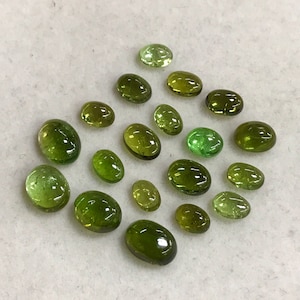 May include: A collection of 18 oval-shaped green gemstones, some with a lighter green hue, arranged on a white surface.