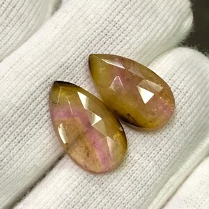 May include: Two faceted, teardrop-shaped gemstones. The gems have a mix of colors, including yellow, brown, and pink. The gems are displayed on a white, textured surface. The gems are likely for jewelry making.