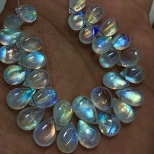 May include: A collection of iridescent, teardrop-shaped moonstone beads. The translucent stones exhibit a play of colour, with flashes of blue, yellow, and orange. The beads are strung on a thin, silver-coloured wire, ideal for jewellery making.